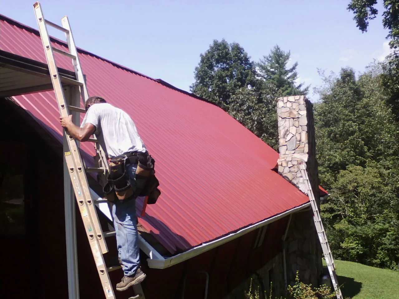 Roofer on a ladder beside a red metal roof and stone chimney — Soffit & Fascia Repair by Roofing Brothers in Cushing
